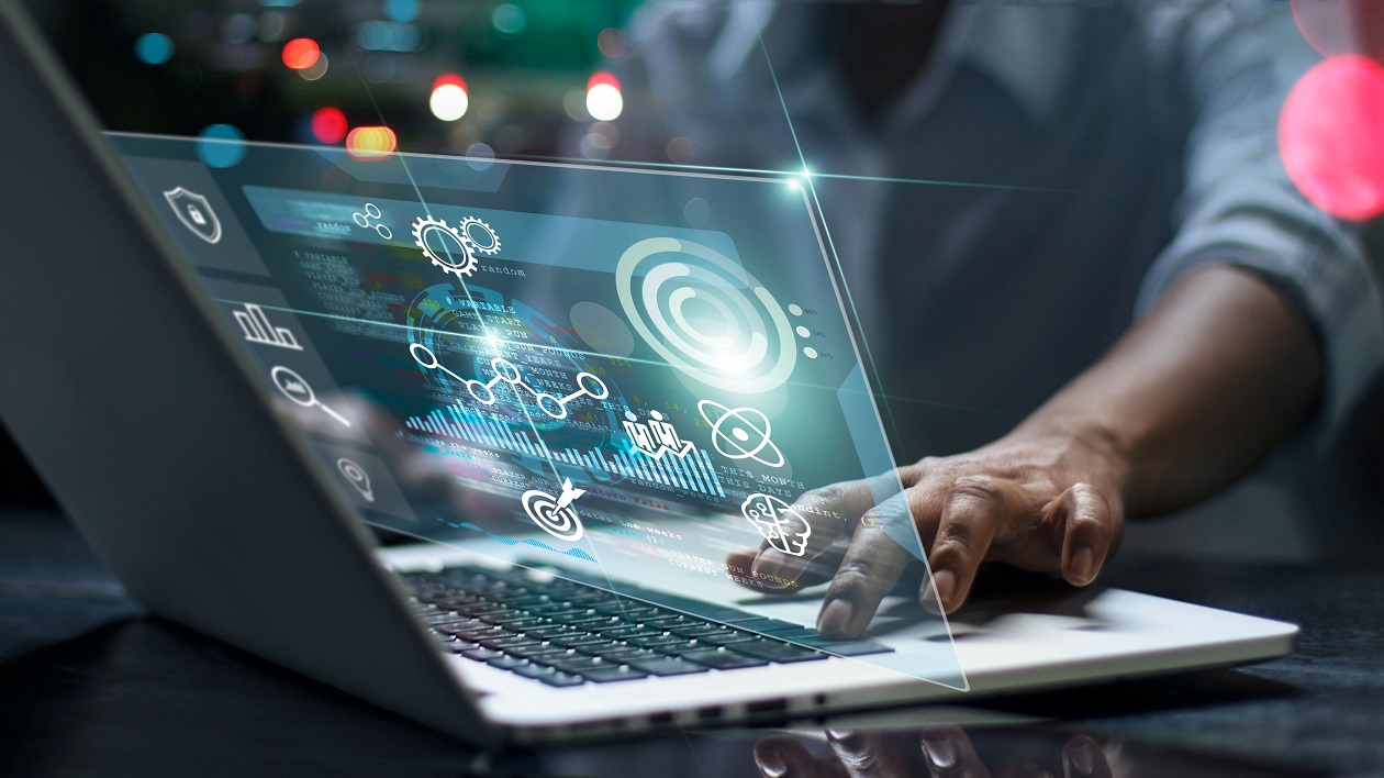 A person working on a laptop with a digital interface projected above the screen, displaying futuristic graphics like data charts, gears, and technological icons, emphasizing innovation, technology, and data analytics.