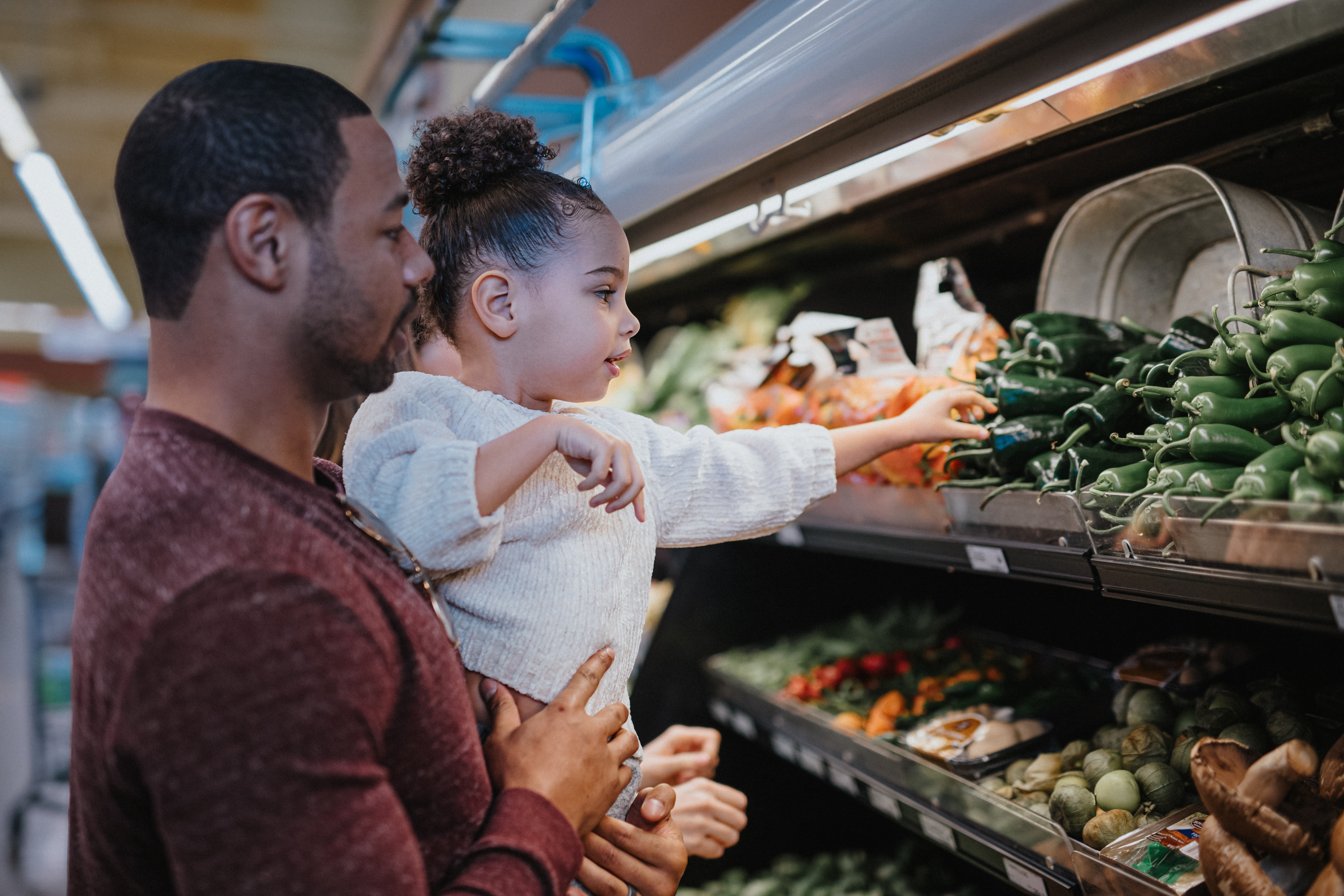 A father holds his young daughter as she reaches for a pepper at the grocery store.
